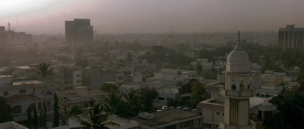 A Mighty Heart (2007) movie still: establishing — A hazy, sprawling cityscape extends to the horizon under a muted, overcast sky. A white minaret…