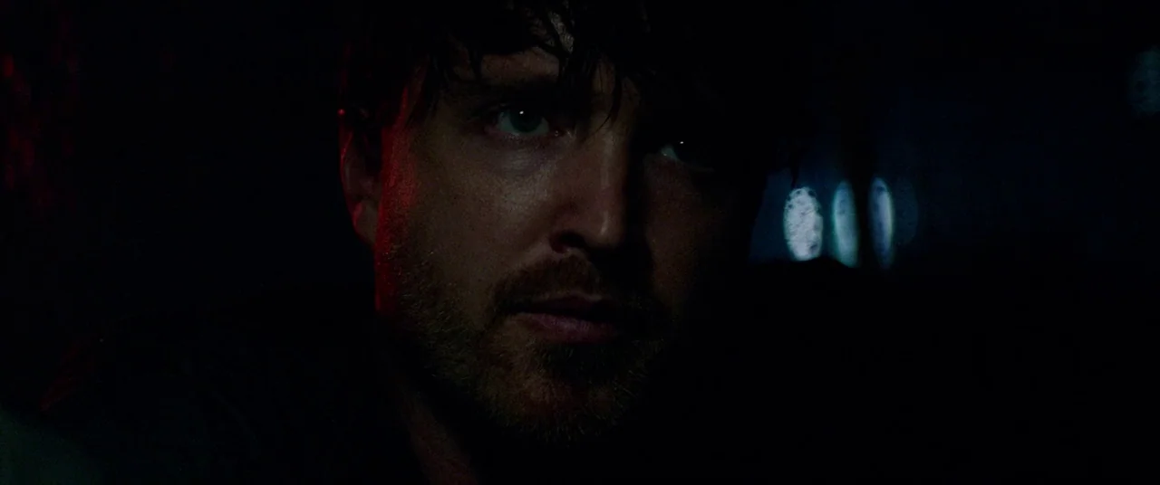 Triple 9 (2016) movie still: closeup — The interior of a dark car at night with faint light reflections visible. A man looks toward the side while…