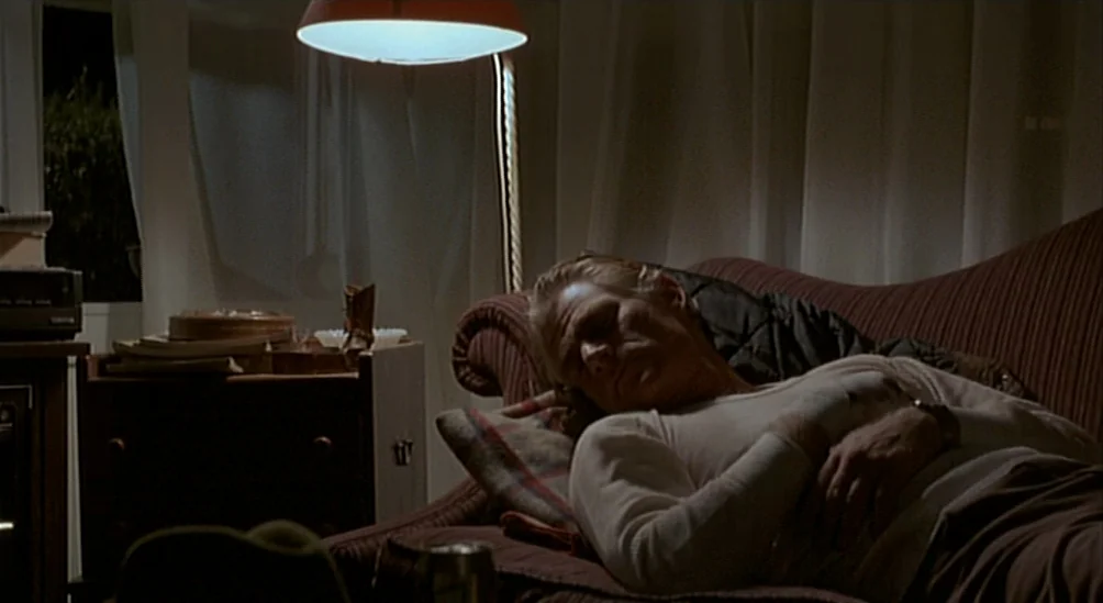 Near Dark (1987) movie still: medium — An interior living room at night features a man sleeping on a striped sofa. A low-hanging pendant light…