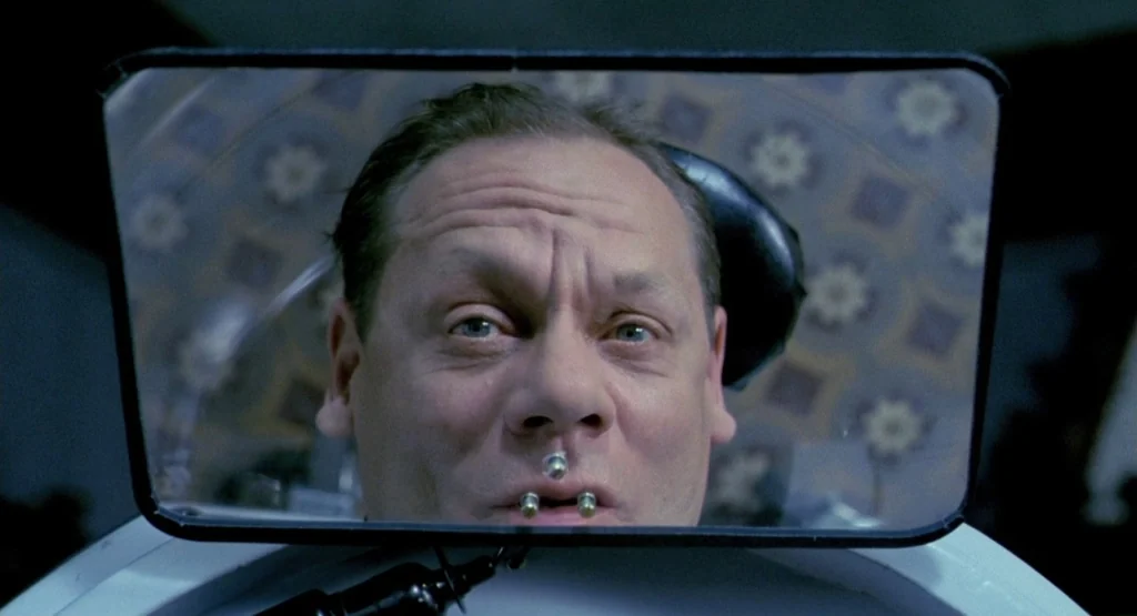 In a Glass Cage (1986) movie still: closeup — A man's face is shown through a small rectangular mirror with patterned wallpaper behind him. He has…