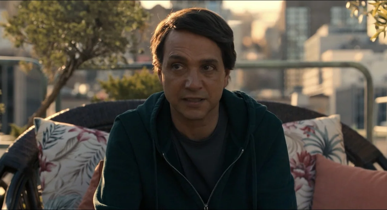 Karate Kid: Legends (2025) movie still: medium — A rooftop terrace with a blurred urban backdrop at golden hour. A middle-aged man sits in a wicker…
