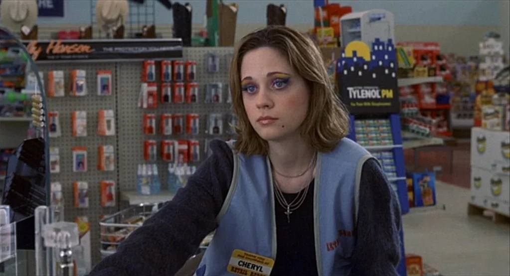 The Good Girl (2002) movie still: medium — A retail store aisle displays various health products on grid wall shelves. A young woman with colorful…