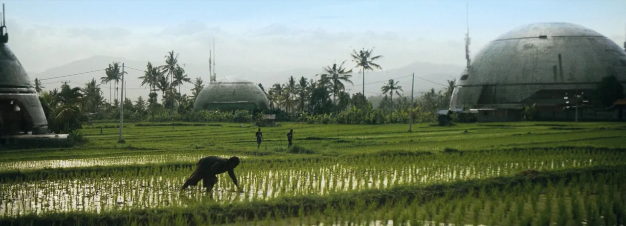 The Creator (2023) movie still: wide — A sprawling green flooded rice field sits under a pale overcast sky. Large metallic domed structures rise…