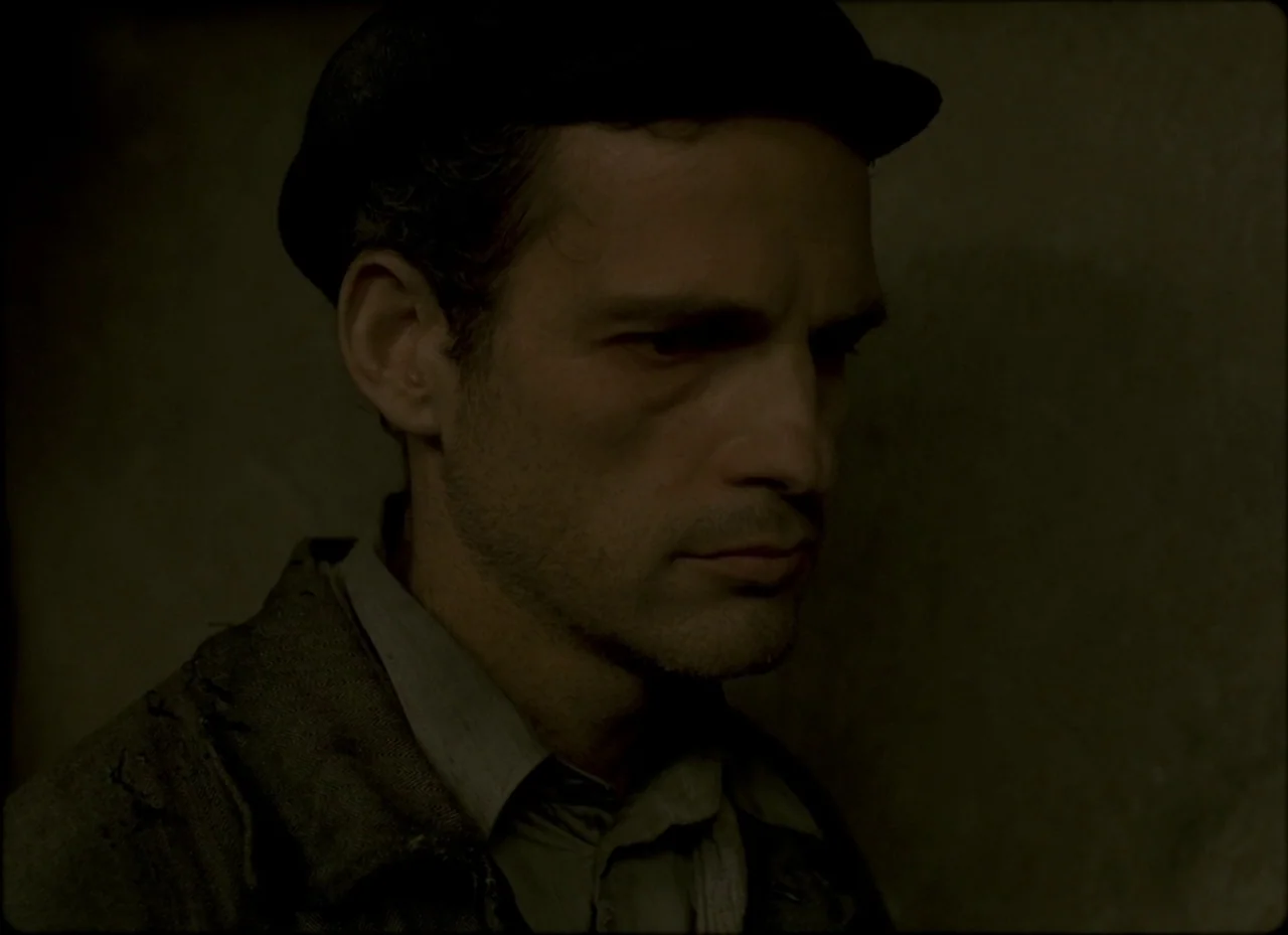 Son of Saul (2015) movie still: closeup — A dimly lit interior space with shadows cast across the wall. A man wears a cap and weathered clothing…