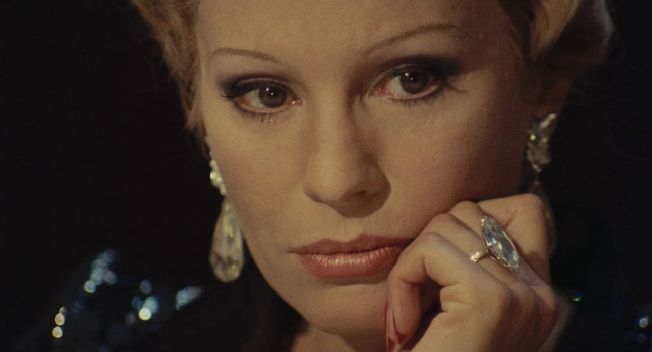 The Damned (1969) movie still: closeup — A close-up of a blonde woman against a dark, featureless background. She rests her chin on her hand,…