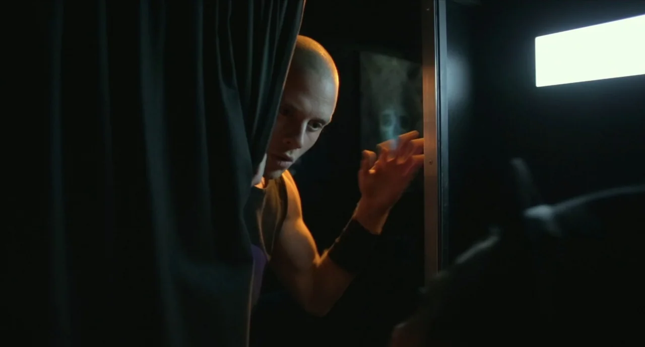 We Are Your Friends (2015) movie still: medium — A dark, cramped interior space features a heavy black curtain and a metal frame. A bald man leans…