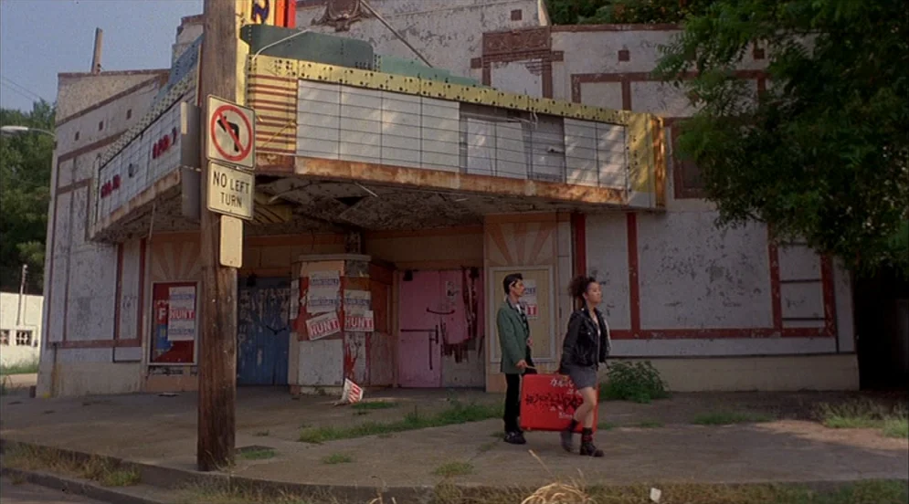 Mystery Train (1989) movie still: wide — A weathered, decaying building with peeling paint stands on a quiet urban sidewalk. A young man and woman…
