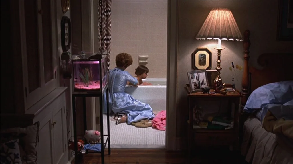 The Exorcist (1973) movie still: medium — A residential bathroom is visible through a doorway from a dimly lit bedroom area. A woman is kneeling on…