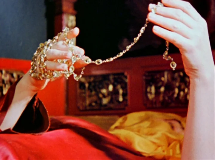 Inauguration of the Pleasure Dome (1954) movie still: closeup — A person lies against a red headboard while holding a multi-strand jeweled necklace.…