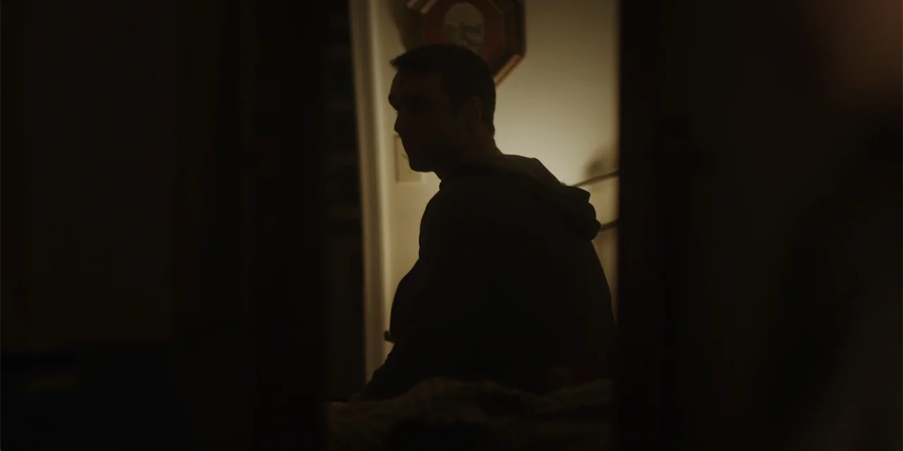 Lobos (2019) movie still: medium — A dark interior room features a man sitting on a bed in low light. The subject is partially silhouetted against a…