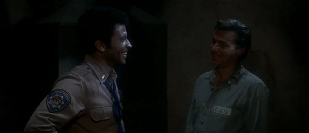 Assault on Precinct 13 (1976) movie still: medium — Two men stand facing each other inside a dimly lit stone room. A uniformed officer smiles at a…