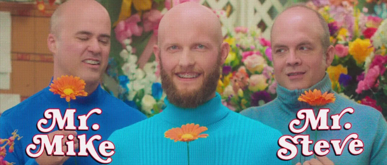 Greener Grass (2019) movie still: medium — Three bald men stand in front of a wall of colorful flowers. Each man holds an orange gerbera daisy in…