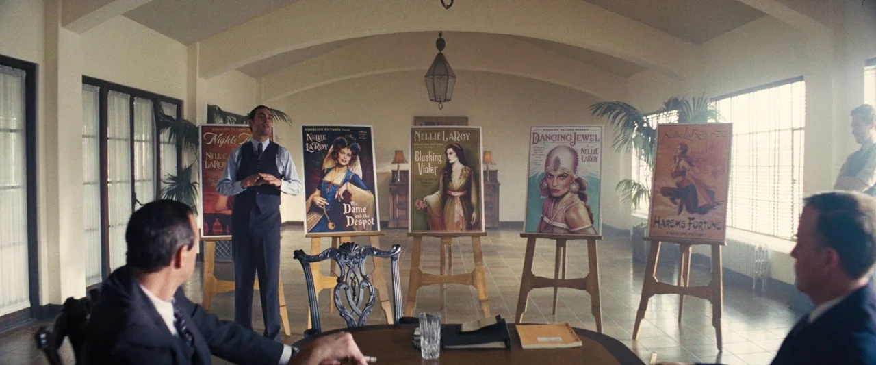 Babylon (2022) movie still: wide — A sunlit meeting room contains a central table and five posters displayed on wooden easels. A man stands beside…