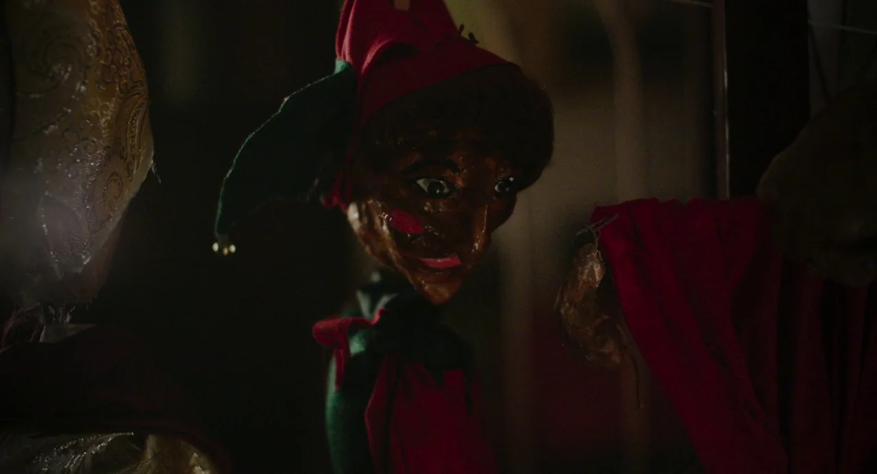All You Need Is Death (2023) movie still: closeup — A dark, dimly lit environment features a small wooden doll with an elf hat. The doll hangs…