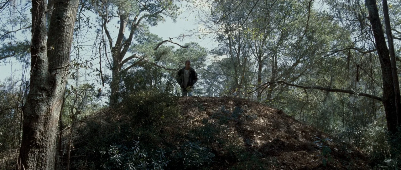 The New Daughter (2009) movie still: wide — A man stands on a dirt rise surrounded by mature deciduous trees and shrubs. The figure in a jacket…