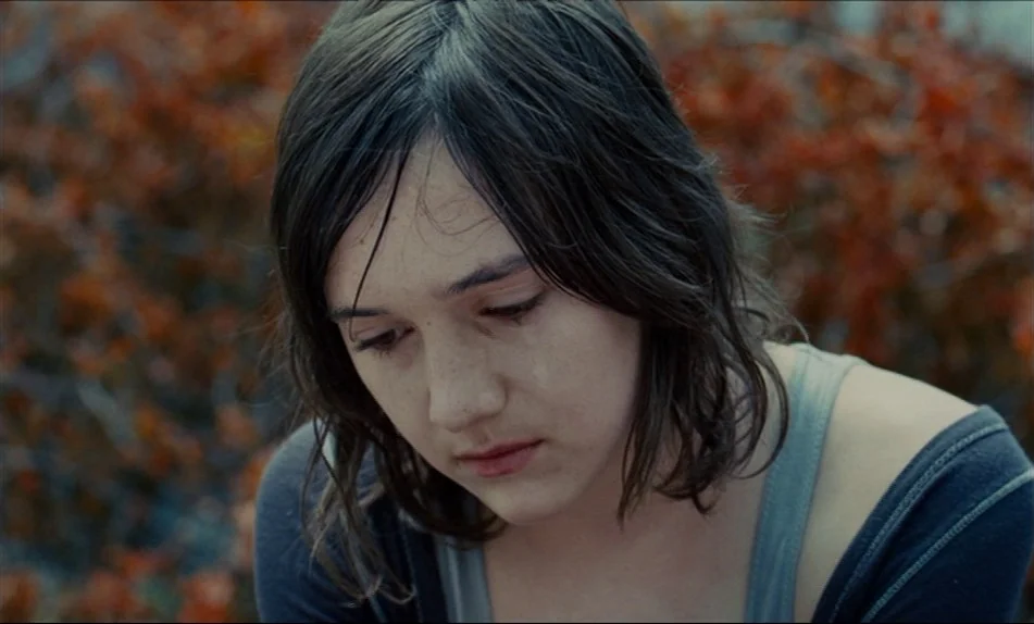 Hadewijch (2009) movie still: closeup — An outdoor park setting features blurred reddish foliage in the out-of-focus background. A young woman with…
