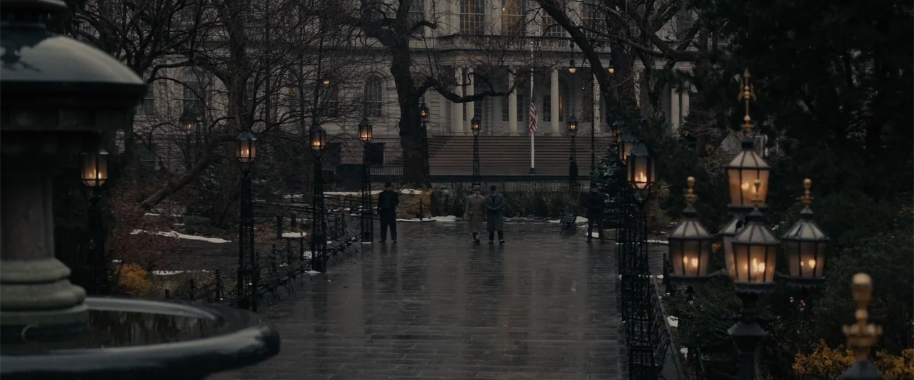 Winter's Tale (2014) movie still: wide — A stone walkway leads through a dimly lit park toward a large administrative building. Three men in…
