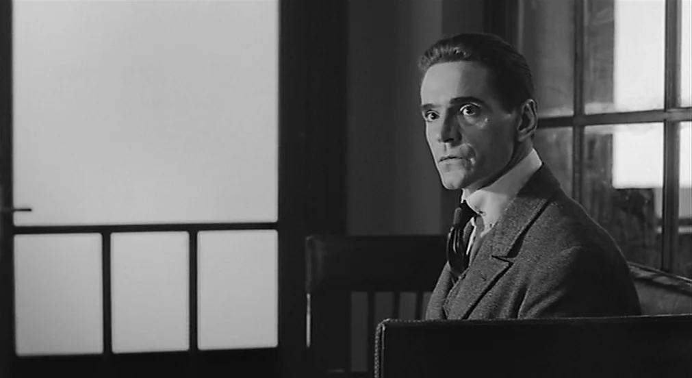 Kafka (1991) movie still: medium — A dark, wood-paneled room features a large window with multiple panes behind the subject. A man wearing a formal…