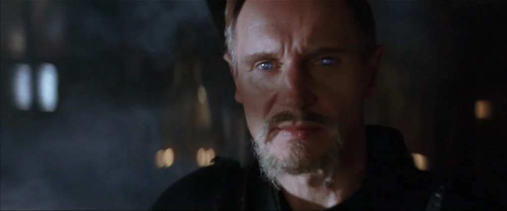 Batman Begins (2005) movie still: closeup — A dark, dimly lit stone interior with indistinct architectural features in the background. A man with a…