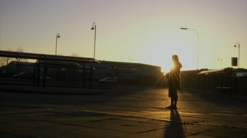 Red Road (2006) movie still: wide — An empty bus terminal at sunrise features covered shelters and lamp posts against a sky. A solitary woman in a…