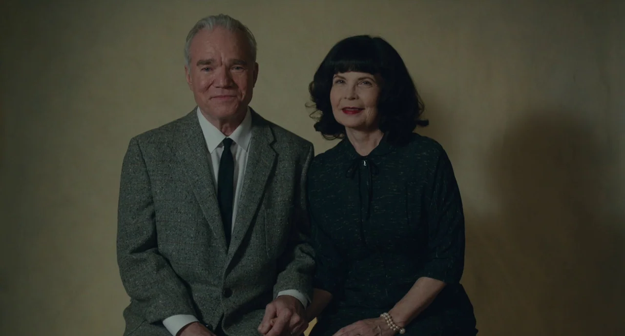 Wildlife (2018) movie still: medium — A man and woman pose for a formal portrait against a plain tan wall. They sit side by side holding hands while…