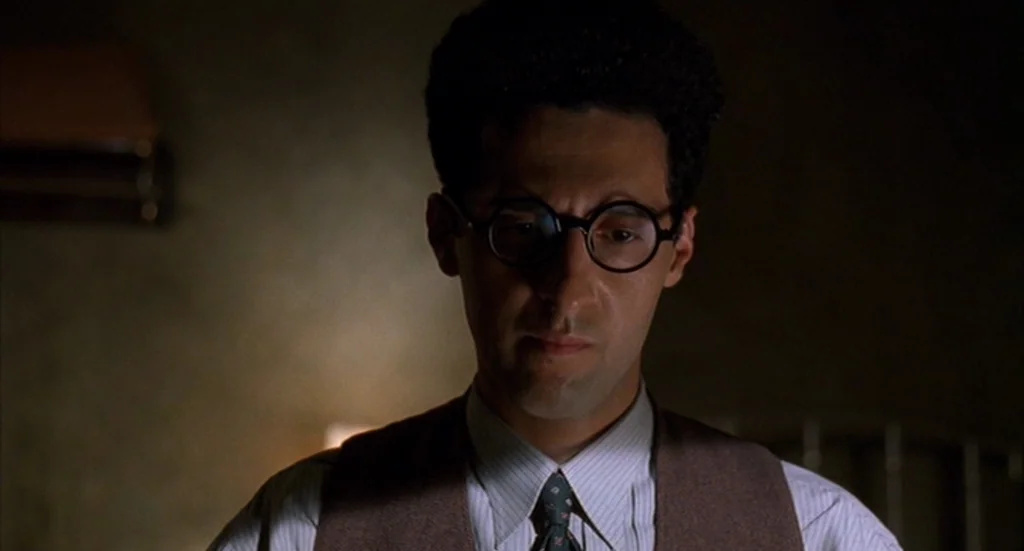 Barton Fink (1991) movie still: closeup — A dimly lit interior space with muted colors. A man in a vest and glasses looks down.