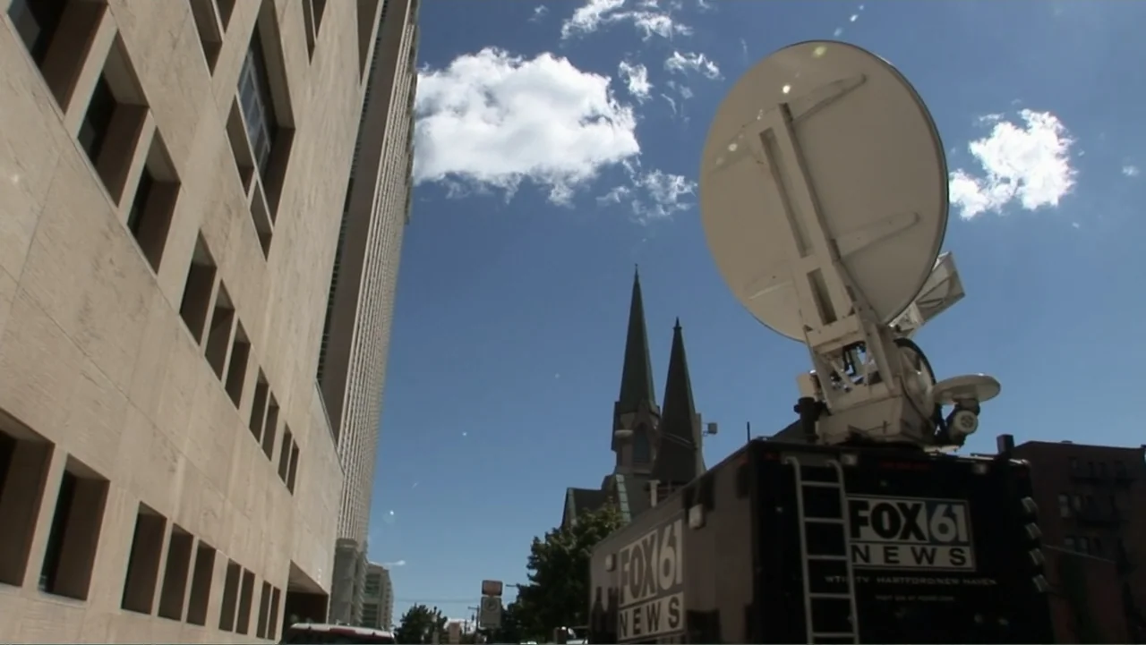 The Cheshire Murders (2013) movie still: wide — A tall modern office building and a church spire occupy a busy city street. A black news truck with…