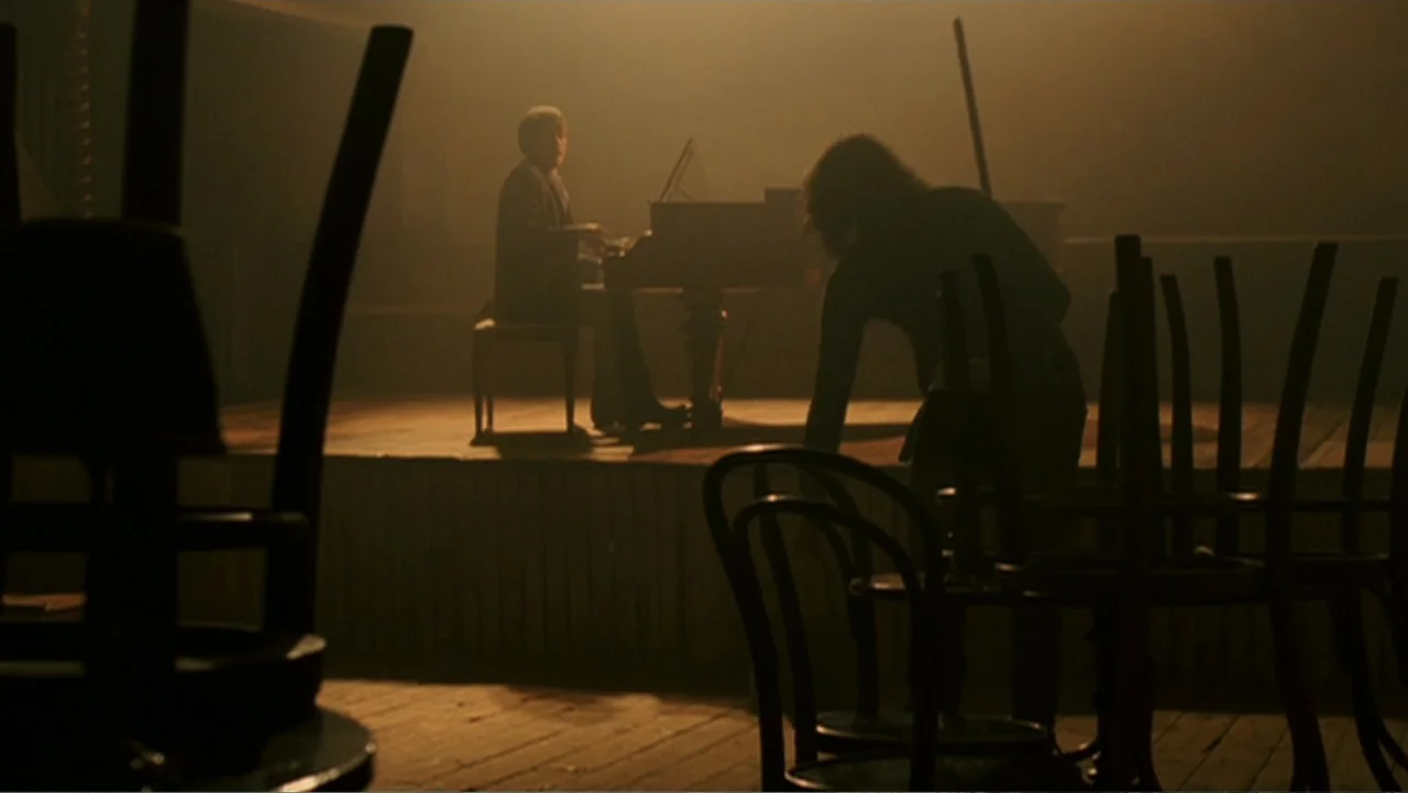Penelope (2007) movie still: medium — A wooden performance space holds a grand piano and several overturned chairs in darkness. A musician plays the…