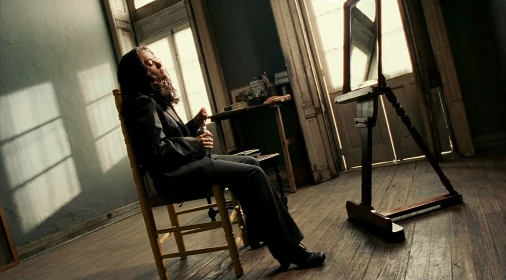 Frida (2002) movie still: fullbody — An artist's space features a wooden chair and an easel set against greenish walls. A woman in a black suit sits…
