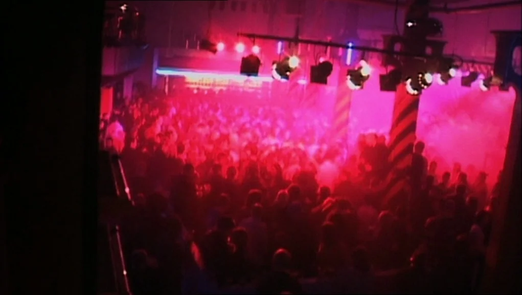 24 Hour Party People movie still: wide — A dense, smoke-filled nightclub floor is illuminated by harsh magenta stage lights. A large crowd of people…