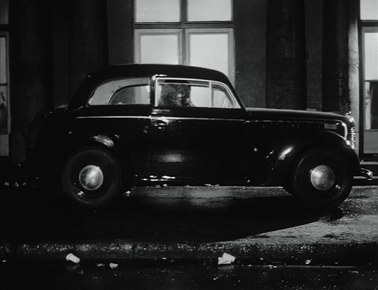 Damnation (1988) movie still: fullbody — A dark vintage sedan is parked on a city street at night. A lone man sits in the driver's seat of the…
