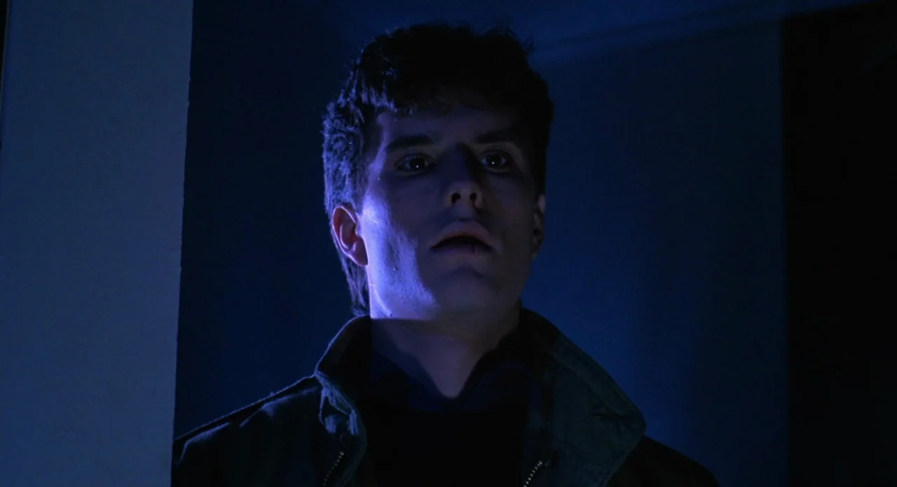 Brain Damage (1988) movie still: closeup — A dark, indistinct indoor space with minimal architectural detail visible in the shadows. A young man…