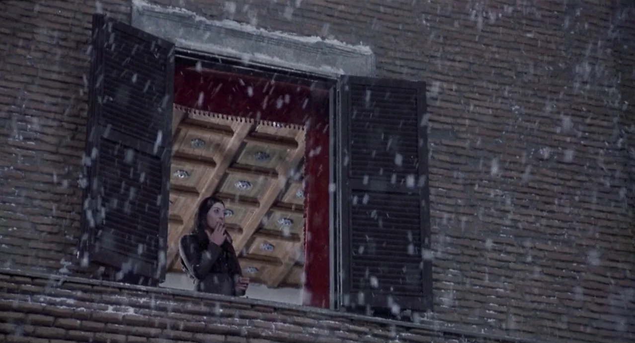 Amarcord (1973) movie still: wide — A brick building facade features an open window with dark wooden shutters during snowfall. A woman stands inside…