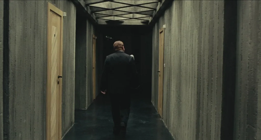 Todo Modo (1976) movie still: fullbody — A long corridor features raw concrete walls with vertical impressions and light wooden doors. A man in a…