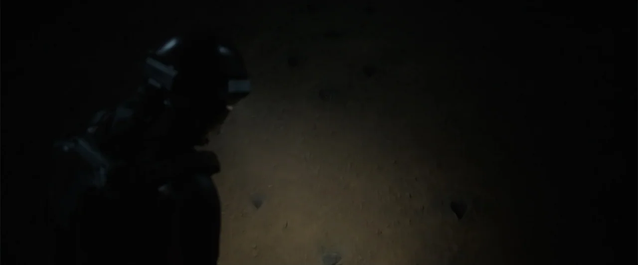 DISTANT (2026) movie still: medium — A dark, sandy exterior at night lit by a concentrated beam of artificial light. A person wearing a tactical…