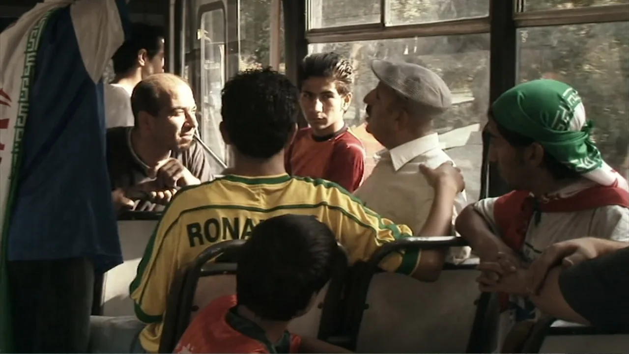 Offside (2015) movie still: medium — The interior of a crowded public bus during daylight hours with passengers seated close. Diverse men in casual…