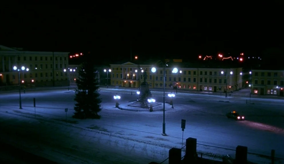 Night on Earth (1991) movie still: establishing — A wide, deserted European square at night is covered in a layer of snow. Several long, yellow,…