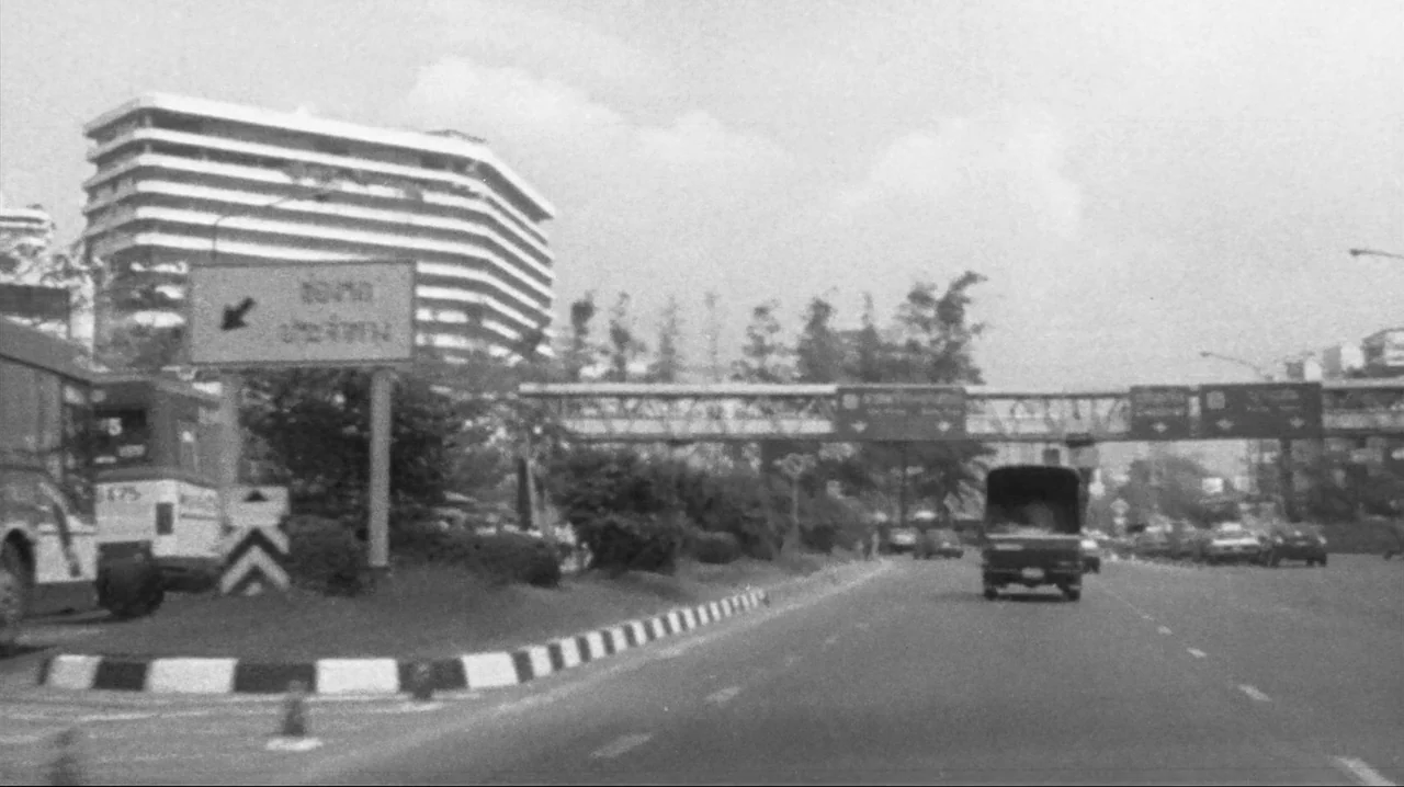 Mysterious Object at Noon (2000) movie still: wide — An urban highway scene in Thailand features a modern high-rise office building nearby. Cars and…