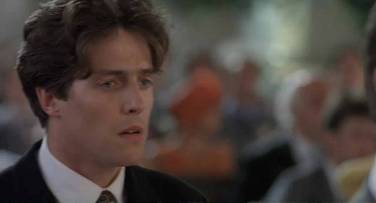 Four Weddings and a Funeral (1994) movie still: closeup — An interior space with a blurred congregation of people in the background. A man in a dark…
