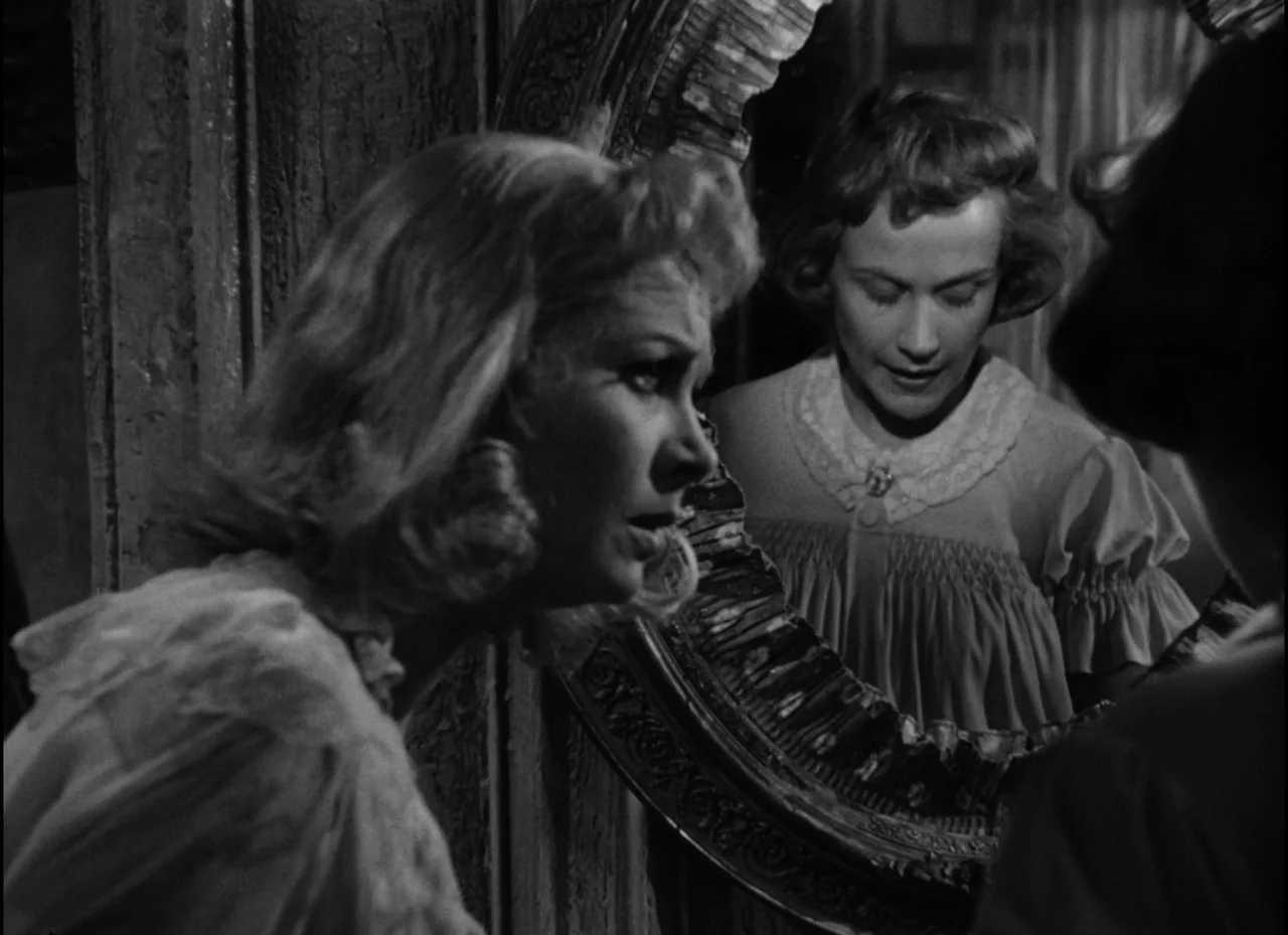 A Streetcar Named Desire (1951) movie still: medium — A woman stands before an ornate antique mirror inside a dimly lit room. She gazes intently at…