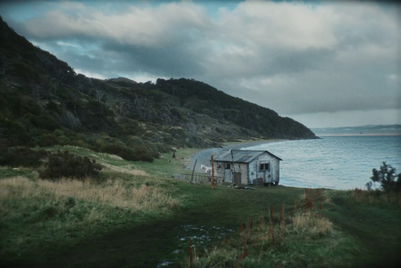 The Settlers (2023) movie still: wide — A wooden cabin stands on a grassy slope beside a turbulent, overcast sea. Rolling hills covered in dark…