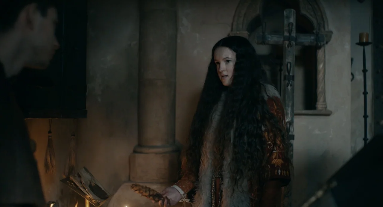 Catherine Called Birdy (2022) movie still: medium — A dark stone chamber features a wooden pillar and an iron cross structure. A young woman stands…