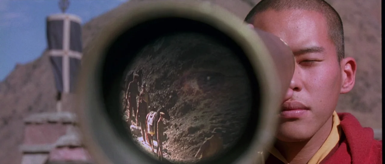 Kundun (1997) movie still: closeup — A person in a red robe uses a telescope in a rocky, mountainous area. The circular lens of the telescope shows…