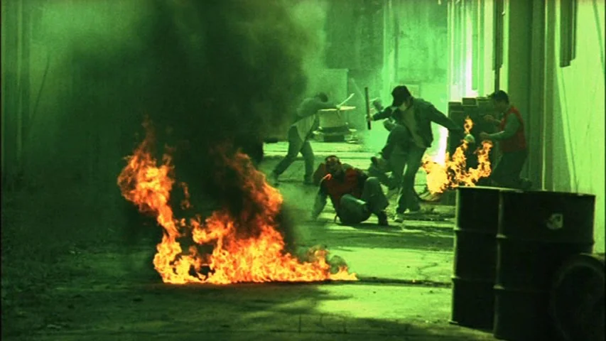 Save The Green Planet! (2003) movie still: wide — A concrete outdoor area is filled with thick black smoke and orange flames. Several men are moving…
