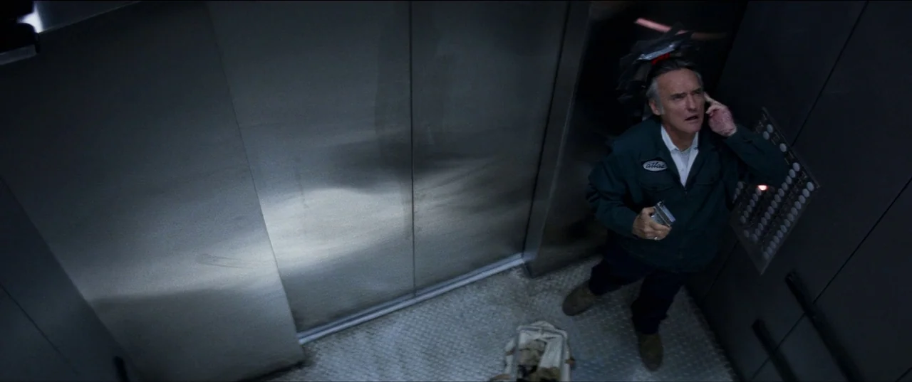 Speed (1994) movie still: fullbody — A modern metallic elevator features brushed steel panels and a diamond plate floor. An older man in a work…