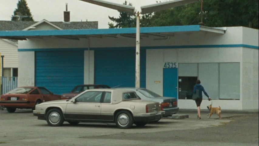 Wendy and Lucy (2008) movie still: wide — A single-story building with a white facade and blue accents stands in a parking lot. A woman walks toward…