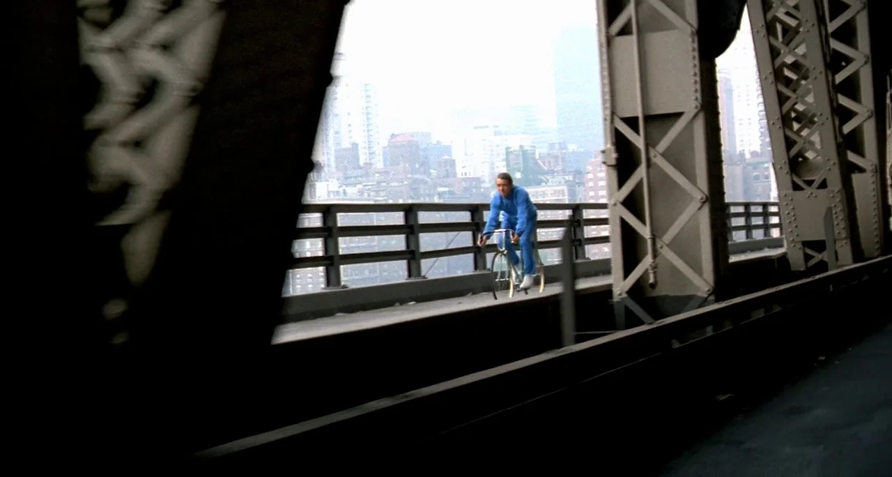 Tenebre (1982) movie still: wide — A metal bridge structure frames a man cycling across a pedestrian path. The urban skyline is visible in the…