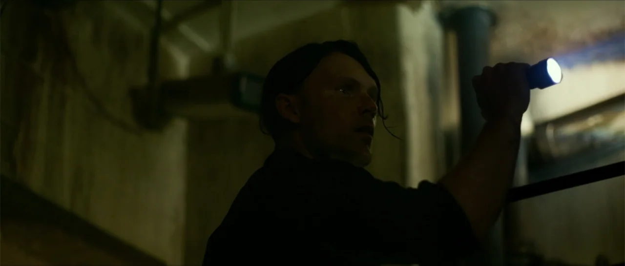 The Vault (2017) movie still: medium — A dimly lit industrial basement featuring exposed concrete walls and metal pipes in the background. A man in…