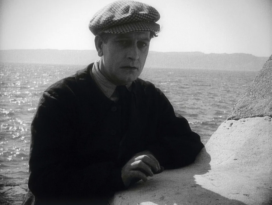 Coeur Fidele (1923) movie still: medium — A man stands outdoors near a body of water with stone structures visible. He rests his arms on a rough…
