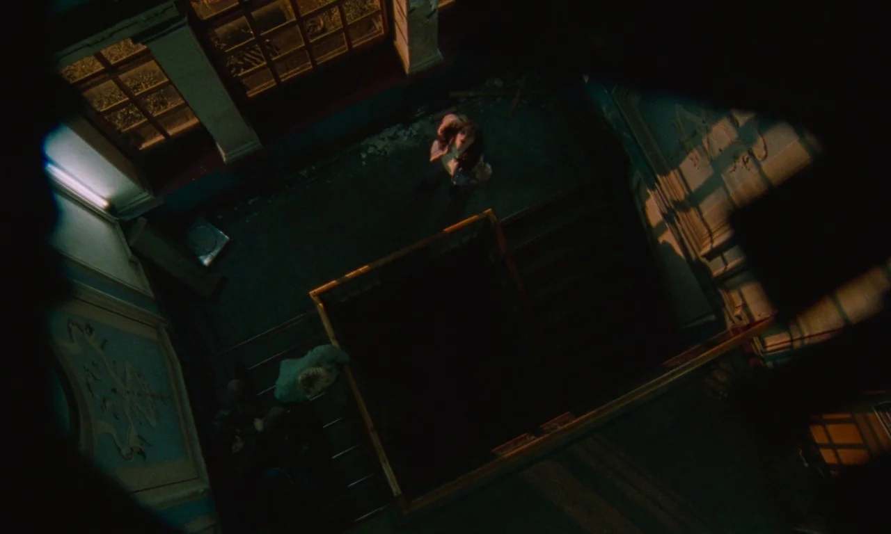Blue Jean (2022) movie still: wide — An ornate dark stairwell with a wooden banister seen from a high-angle overhead perspective. A young woman…